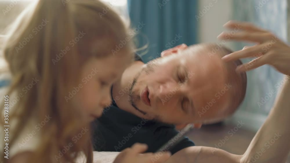 CU: young father tired with helping cute little daughter with ponytails and smartphone do homework lie on floor in light room at quarantine time CU: young father tired with helping cute little daughter with ponytails and smartphone do homework lie on floor in light room at quarantine time