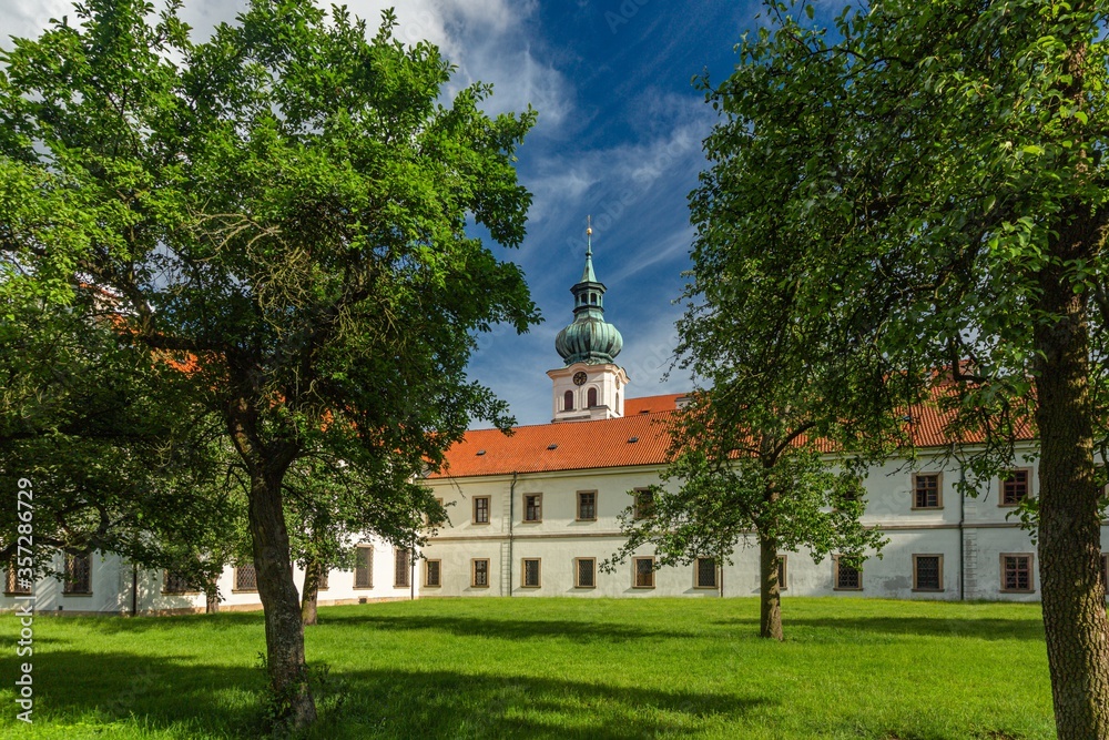 Fototapeta premium Brevnov, Prague / Czech Republic - June 11 2020: View of the first male monastery in Bohemia standing in a park with green trees on a sunny summer day, blue sky with white clouds.