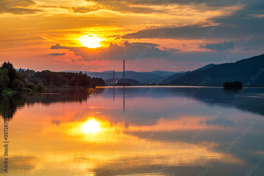 Industrial landscape with chimney constructions reflected on the water surface of the dam at sunset. Zilina dam in the north of Slovakia, Europe.