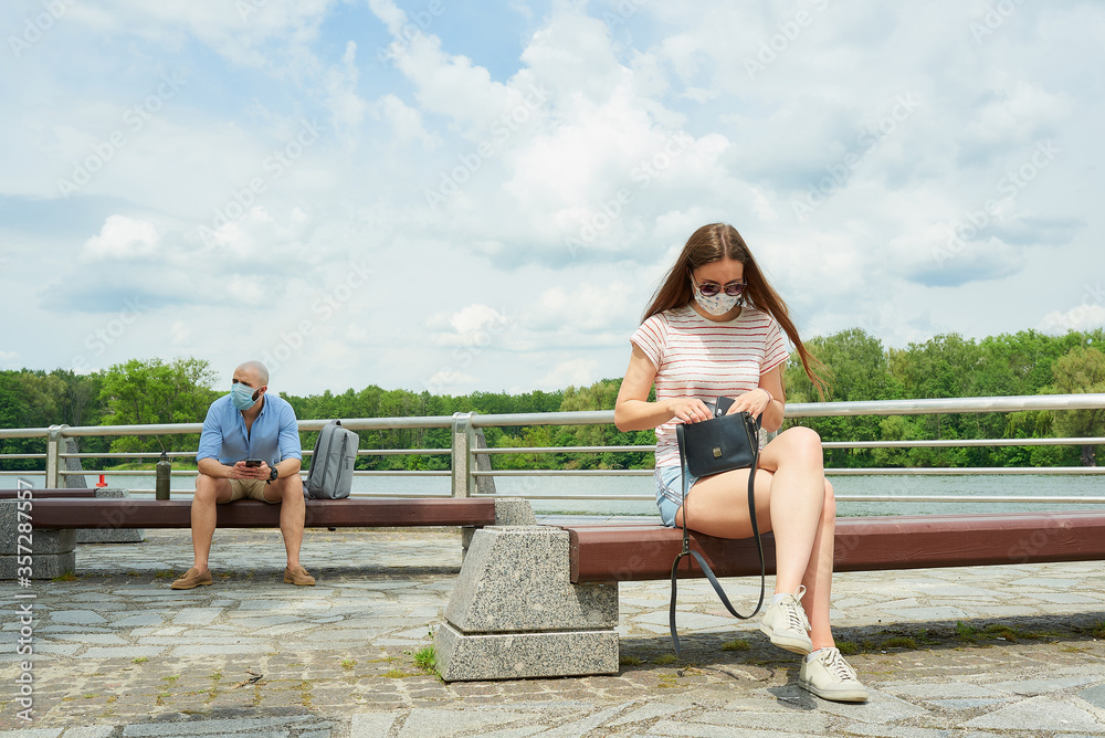 A woman in a face mask sitting on a bench keeping a social distance with a man sitting on another bench on the promenade. Girl searching for phone in bag and guy using smartphone in social distancing.