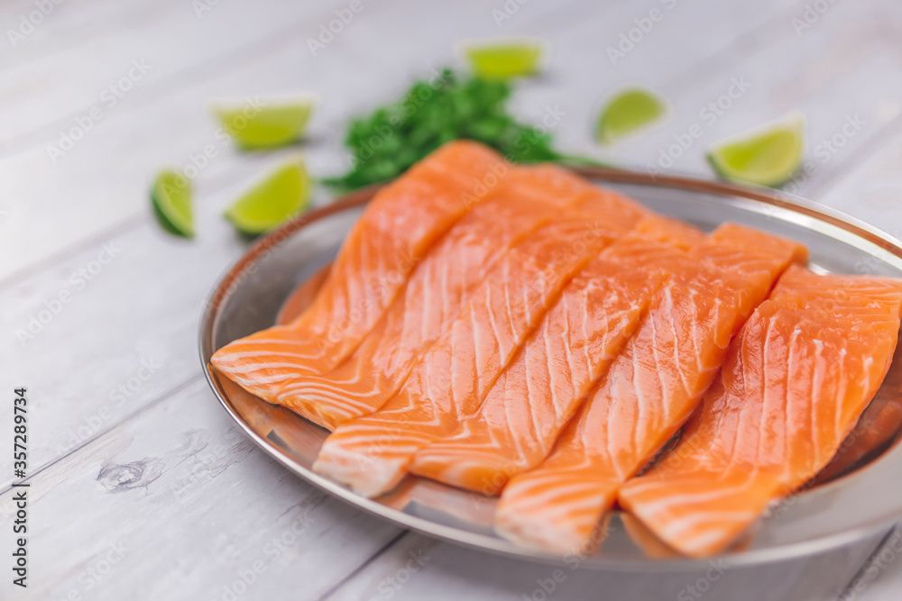 Raw vibrant salmon fillet in a serving plate with herbs, creamy sauce made from butter, garlic, and vegetables. Salmon with wedge lime on the side on a white wooden table. Raw vibrant salmon fillet in a serving plate with herbs, creamy sauce made from butter, garlic, and vegetables. Salmon with wedge lime on the side on a white wooden table.