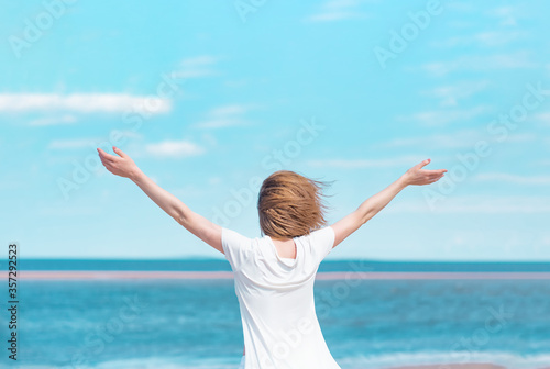 young woman in white clothes stands on the background of the sea, raising her hands to the sky