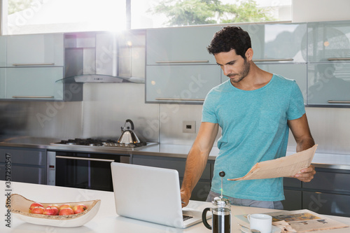 Man with newspaper using laptop in kitchen