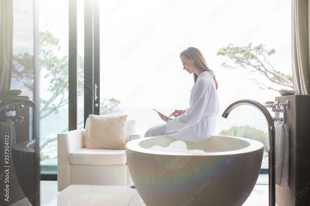 Woman wearing white bathrobe sitting on edge of modern bathtub using digital tablet Woman wearing white bathrobe sitting on edge of modern bathtub using digital tablet