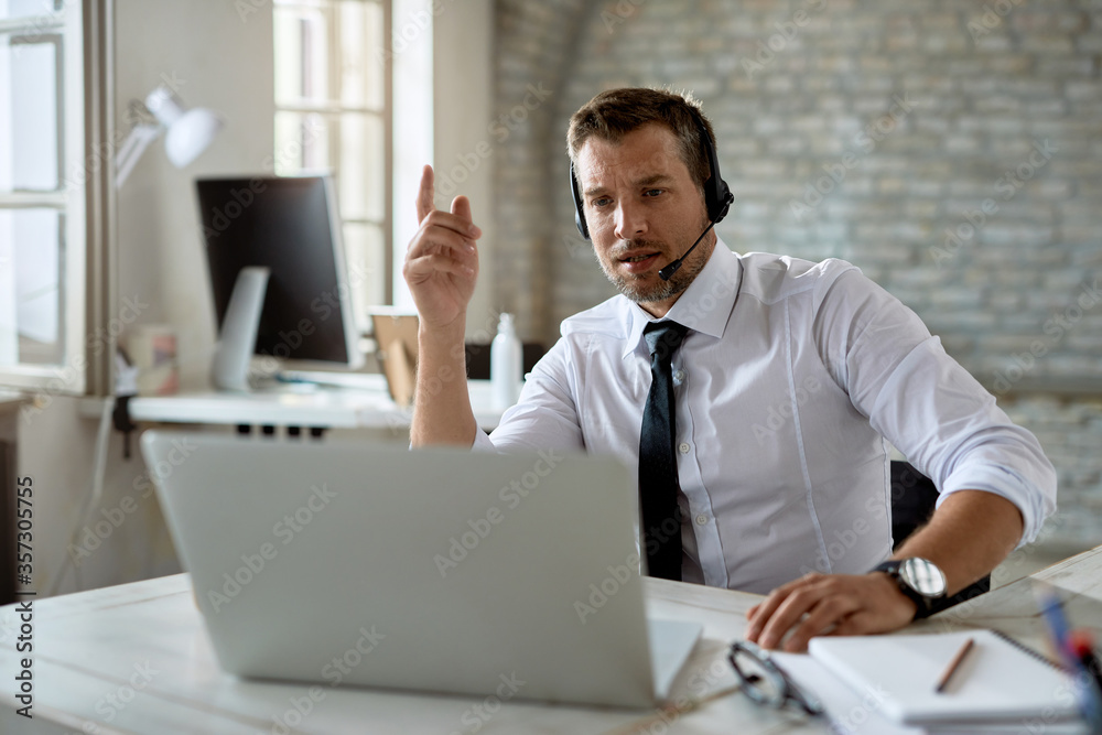 Mid adult businessman wearing headphones while having video call over laptop in the office. Mid adult businessman wearing headphones while having video call over laptop in the office.