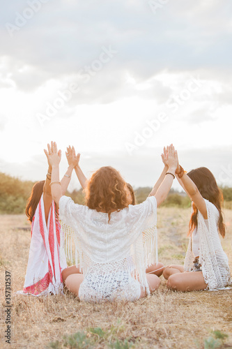 Boho women sitting in circle with arms raised connected in rural field