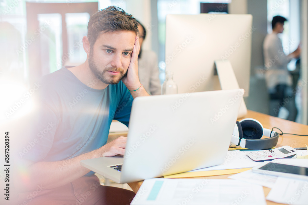Serious creative businessman using laptop with head in hands in office Serious creative businessman using laptop with head in hands in office