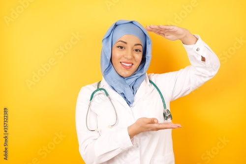 Young beautiful doctor muslim woman wearing a medical uniform and hiyab over isolated background gesturing with hands showing big and large size sign, measure symbol. Smiling looking at the camera.  photo