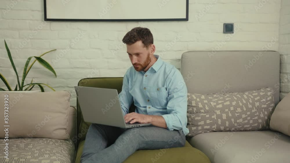 Happy man using laptop typing look at camera waving his hand wants to show something sitting on couch in modern living room at home internet business computer close up slow motion Happy man using laptop typing look at camera waving his hand wants to show something sitting on couch in modern living room at home internet business computer close up slow motion