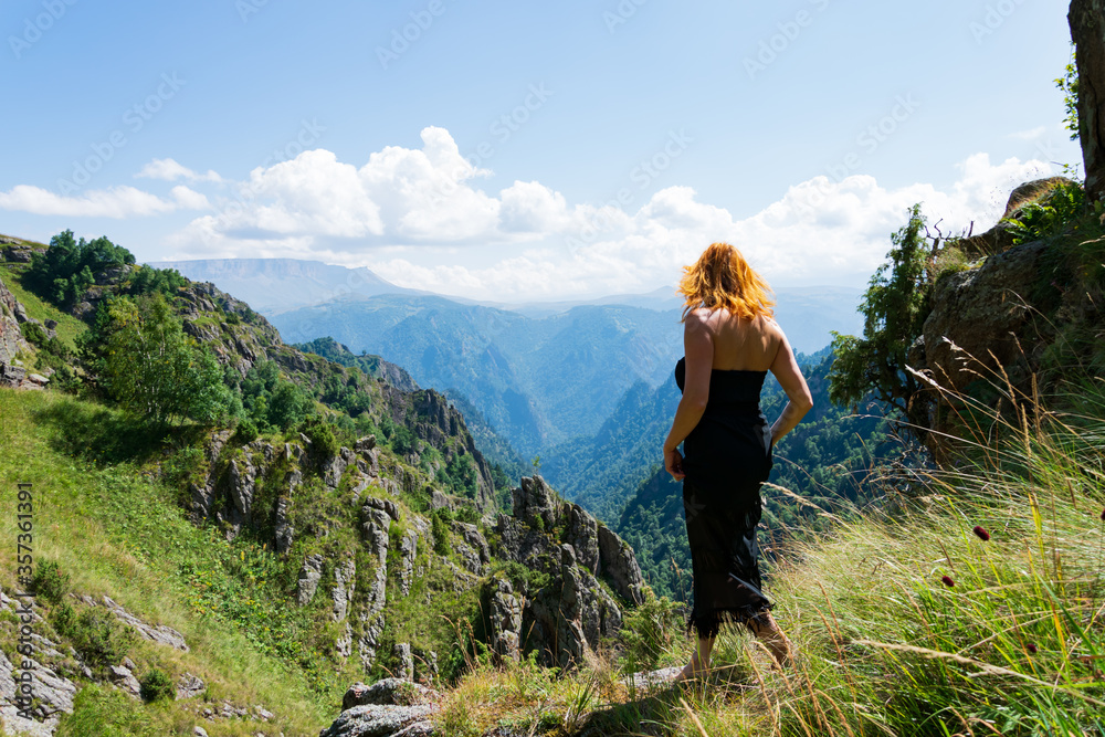 A beautiful girl in a black dress with a cleavage, a European woman with red hair, walking barefoot along a mountain range.