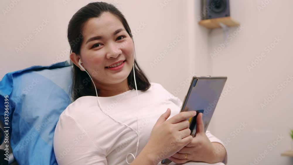Beautiful asian woman look camera and smile relaxing and listening to music by tablet with headphones on the bed at home lifestyle and pleasure concept. Beautiful asian woman look camera and smile relaxing and listening to music by tablet with headphones on the bed at home lifestyle and pleasure concept.