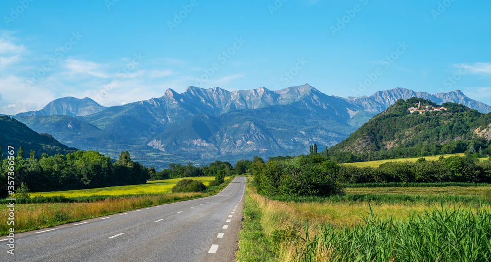 A long straight road leading towards a mountains in France. Amazing bright colorful spring and summer landscape. Yellow fields of flowering rape and blue sky with clouds. Natural landscape background.