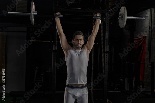 Selective focus of good looking young Caucasian sport man doing exercise by weight lifting in a dark gym. White strong man got sweat while lifting weight in gym with shadow background. Body building