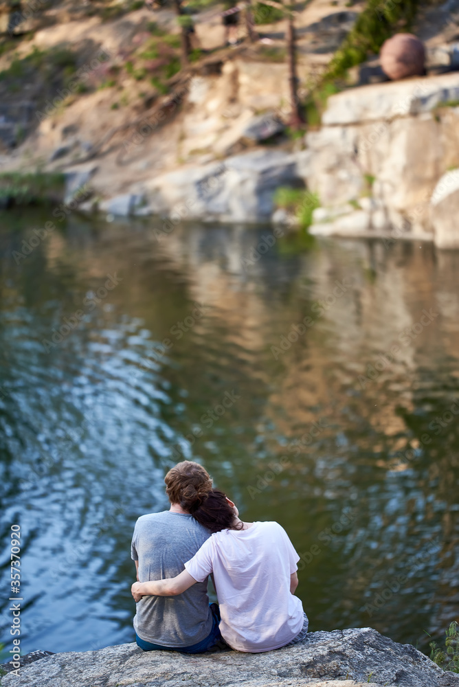 The guy and the girl are sitting on a cliff above the river and looking in front. rear view. loving couple. beautiful vertical shot The guy and the girl are sitting on a cliff above the river and looking in front. rear view. loving couple. beautiful vertical shot