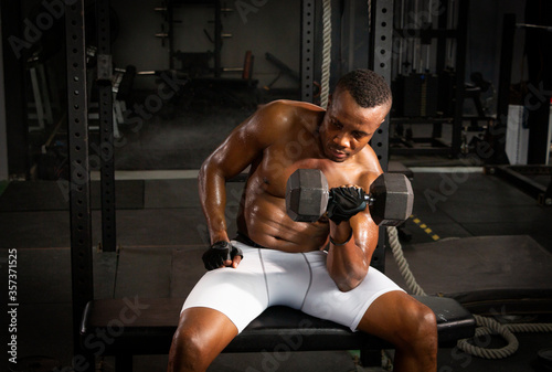 Selective focus of young African athlete sitting on a chair using dumbbell to build his biceps in a dark gym alone. African American man workouts by himself with dumbbell in shadow tone background
