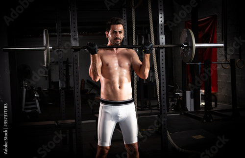 Selective focus of young good looking Caucasian in a position to lift barbell in dark gym by himself. White muscle man workouts with barbell alone to build his body. Smoke background