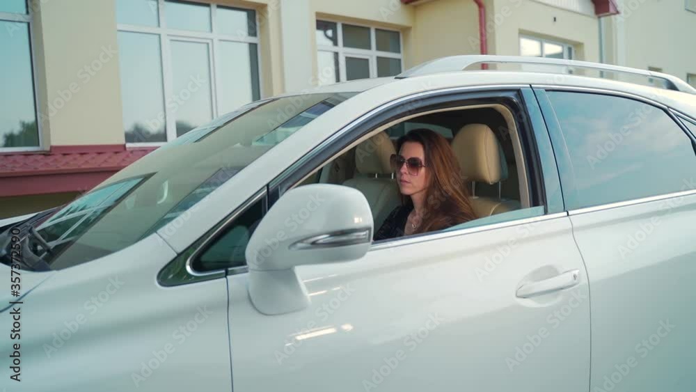 Portrait of a young woman in sunglasses rides in a luxury white car with an open window. The car is in motion. Attractive female with long hair on a building background Portrait of a young woman in sunglasses rides in a luxury white car with an open window. The car is in motion. Attractive female with long hair on a building background