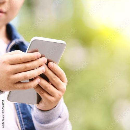 Young school boy texting outdoor. Male person using smartphone. Little hands with cell phone. Green bokeh street background. © elenavolf