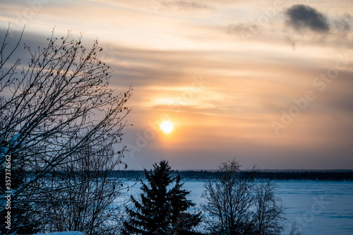 Landscape of sundown over a frozen river and snowy forest in the background.
