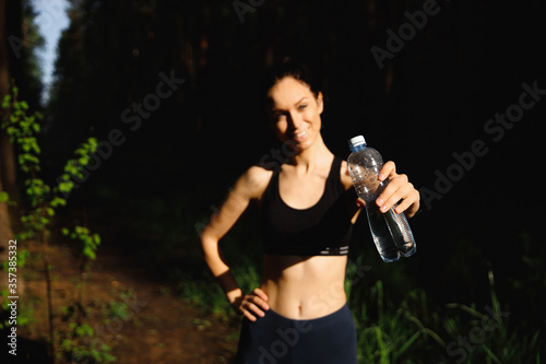 A beautiful woman at arm's length holds a bottle of water in the spruce forest nature for rest after running. Selective focus