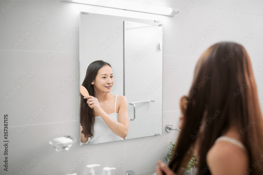 Beautiful young woman in white undershirt is combing her hair and smiling while looking into the mirror in bathroom Beautiful young woman in white undershirt is combing her hair and smiling while looking into the mirror in bathroom
