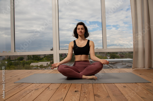 Young woman with short black hair practicing yoga lotus position in the apartment. She is sitting on the wooden floor behind her cloudy sky