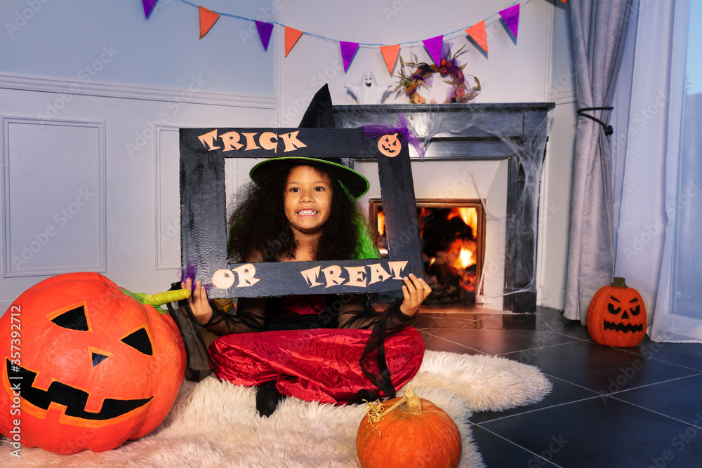 Fototapeta premium Girl with trick or treat frame in Halloween dress sit at home near fireplace and big orange pumpkins