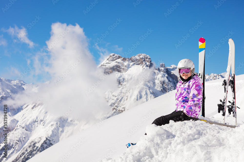 Happy girl in ski outfit, helmet and mask sit in the snow on mountain top look at camera
