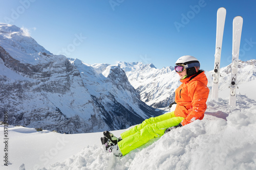 Teenage girl in orange bright sport outfit with ski over high mountain range peaks and blue sky