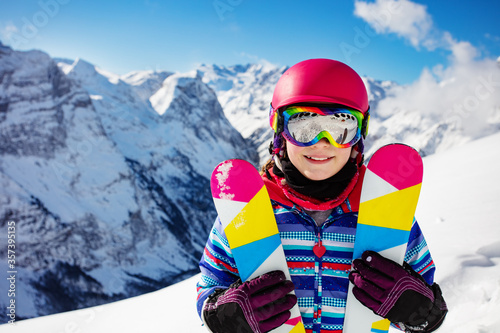 Close-up portrait of cute girl in pink helmet and color hold crossed pair of ski smiling, goggles over high mountain