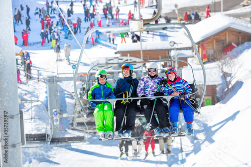 Four kids boy and girls sit together on chairlift lifting on the mountain with crowded resort on background