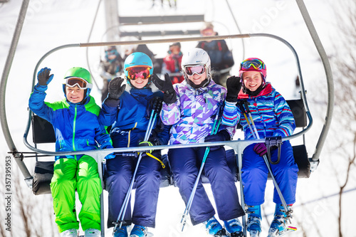 Group of four children sit on the chair lift and wave hands going to the mountain top on alpine ski resort