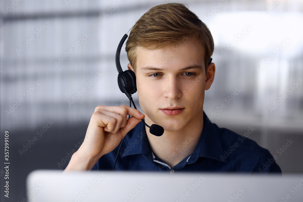 Young blond businessman using headset and computer at work. Startup business means working hard and out of time for success achievement Young blond businessman using headset and computer at work. Startup business means working hard and out of time for success achievement