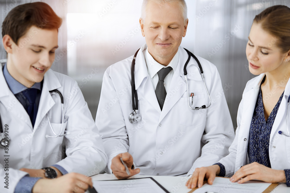 Doctors colleagues and elderly therapist discussing current disease therapy while sitting at working place in sunny clinic. Teamwork in medicine Doctors colleagues and elderly therapist discussing current disease therapy while sitting at working place in sunny clinic. Teamwork in medicine