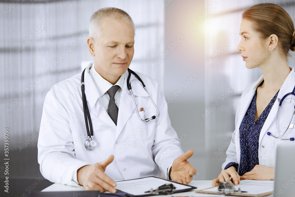 Elderly doctor and his female colleague discussing current disease therapy while sitting at working place in sunny clinic. Teamwork in medicine