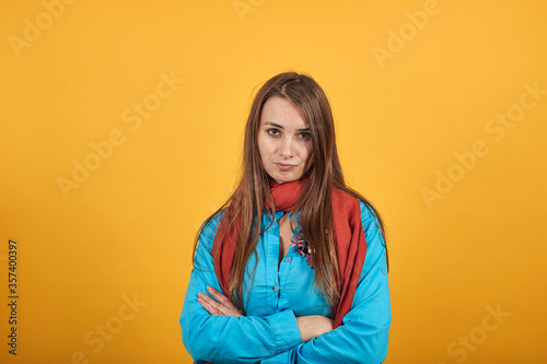 Standing and cross her arms on chest. Young attractive woman with brown hair, eyes, decoration on blue shirt, yellow background © Petro