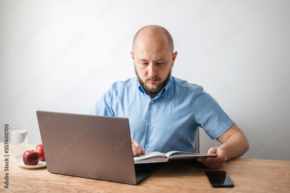 Bald young man with beard reading encyclopedia book and working online on his laptop at the wooden table in home office, white wall as background. Business, freelance or study concept Bald young man with beard reading encyclopedia book and working online on his laptop at the wooden table in home office, white wall as background. Business, freelance or study concept