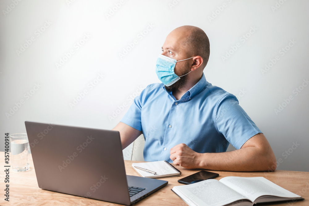 Bald business man with beard looking at the side, wearing surgical medical mask on face as protection against coronavirus, covid-19, modern laptop and book on desk at office, white wall as background Bald business man with beard looking at the side, wearing surgical medical mask on face as protection against coronavirus, covid-19, modern laptop and book on desk at office, white wall as background