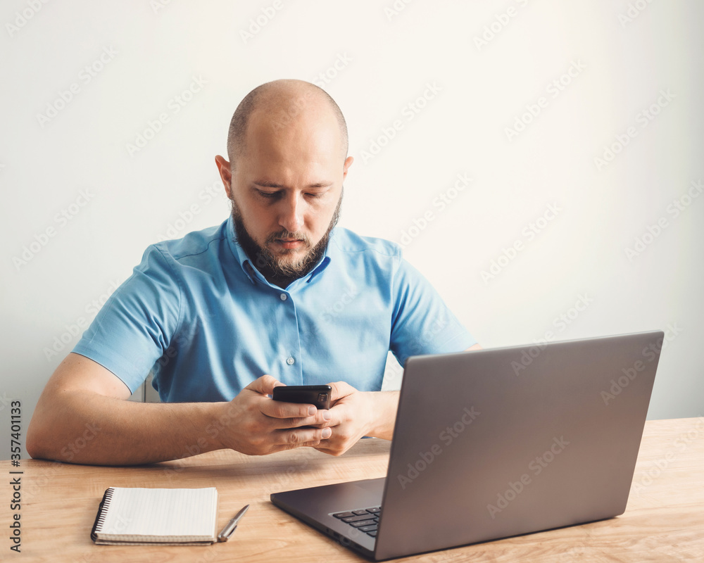 Bald business man with beard uses smartphone and working online, modern laptop on wooden table in home office, white wall as background, business, freelance or study concept Bald business man with beard uses smartphone and working online, modern laptop on wooden table in home office, white wall as background, business, freelance or study concept