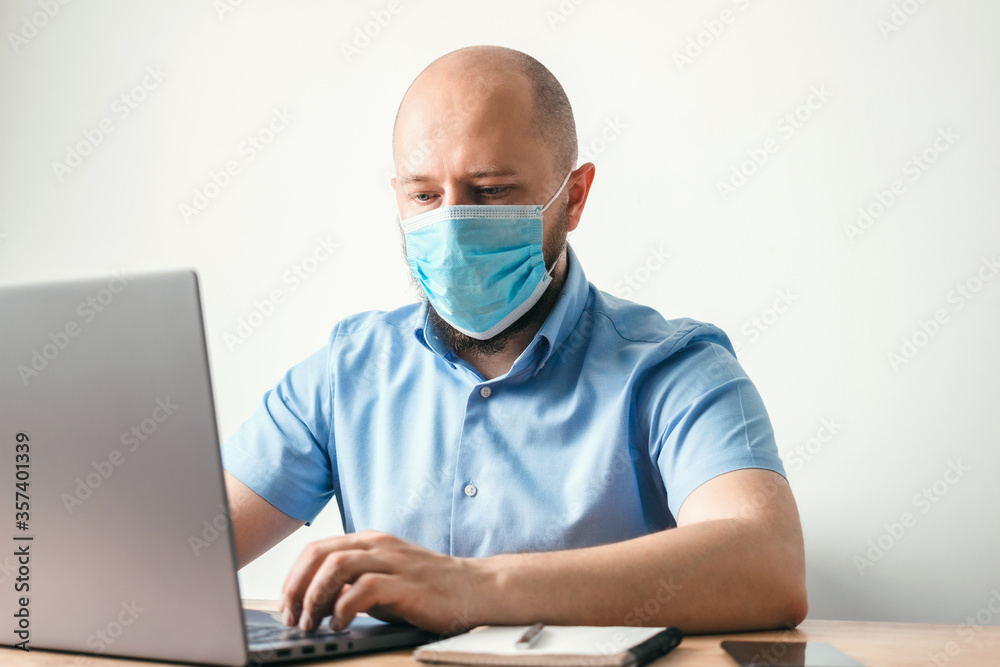 Bald business man with beard wearing surgical medical mask on face as protection against coronavirus, covid-19, working or studying online on laptop at office, social isolation, quarantine concept Bald business man with beard wearing surgical medical mask on face as protection against coronavirus, covid-19, working or studying online on laptop at office, social isolation, quarantine concept