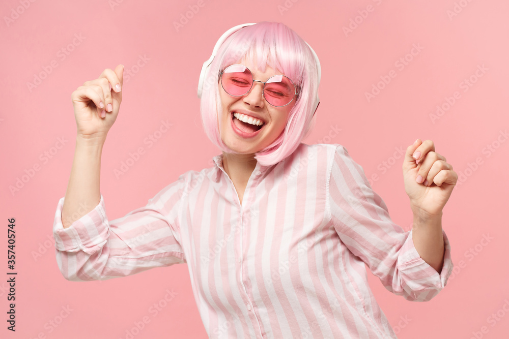 Young happy laughing girl wearing shirt, colored wig and glasses, listening to music in wireless headphones and dancing, isolated on pink background Young happy laughing girl wearing shirt, colored wig and glasses, listening to music in wireless headphones and dancing, isolated on pink background