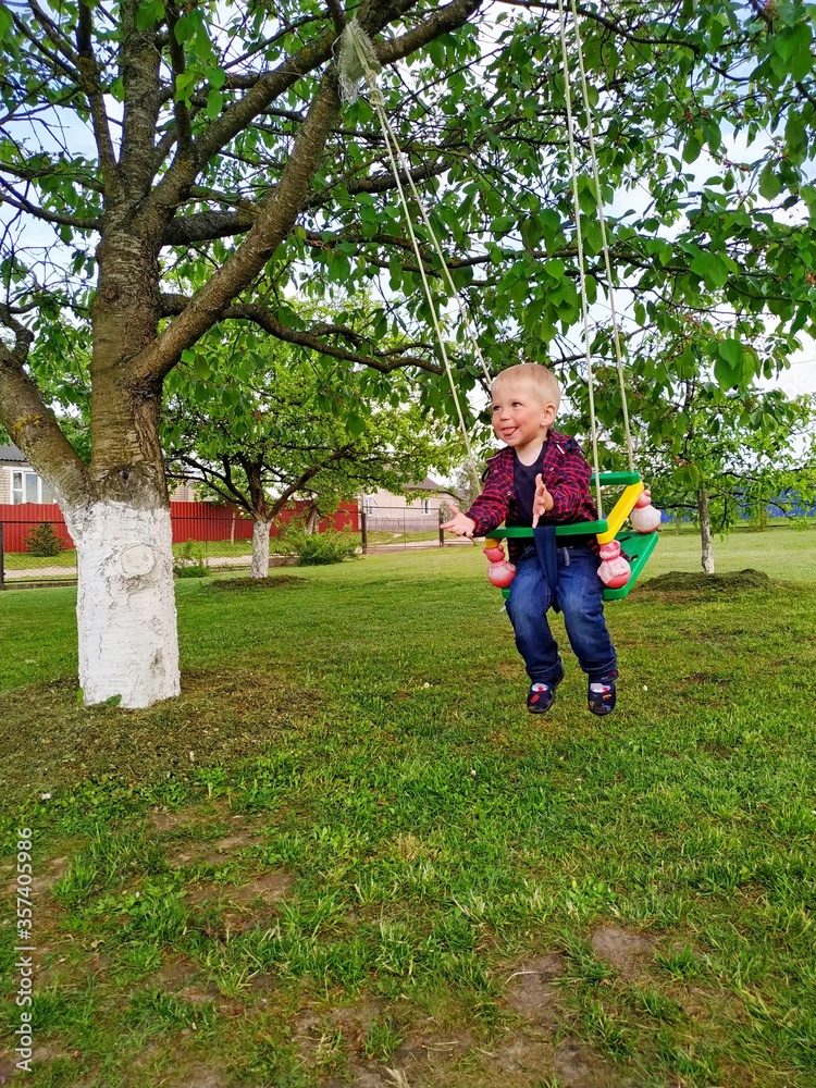 Fototapeta premium joyful kid dressed in a red plaid shirt and blue jeans swinging on a swing on the backyard. Happy moments