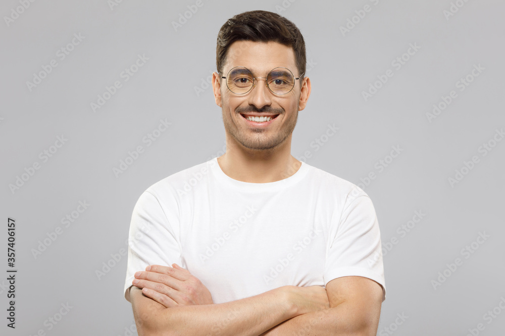 Young handsome smiling man wearing white t-shirt and eyeglasses, standing with arms crossed, feeling confident as professional, isolated on gray background Young handsome smiling man wearing white t-shirt and eyeglasses, standing with arms crossed, feeling confident as professional, isolated on gray background
