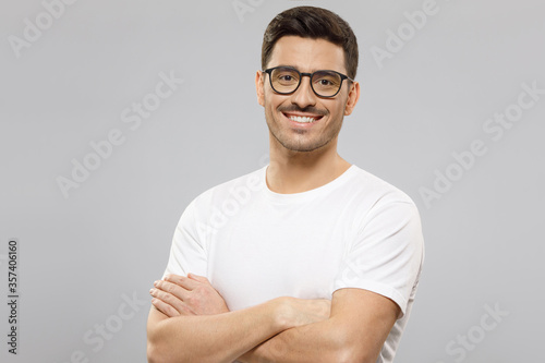 Young confident man wearing white t-shirt and glasses, standing with crossed arms, isolated on gray background