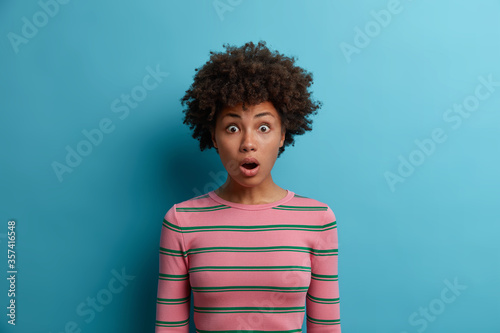 Emotional stupefied dark skinned woman looks in disbelief at camera, keeps jaw dropped, being astonished to hear shocking news, stands astounded against blue background, wears striped jumper