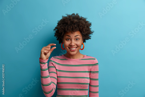 Some more, please. Cheerful smiling African American woman gestures small size with fingers, asks for little bit time, measures too small object, shows something minimum dressed casually stands indoor