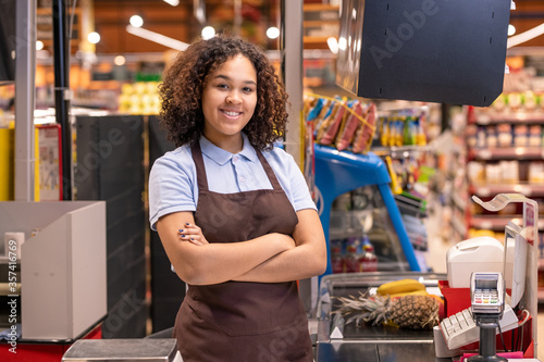 Girl in apron standing by cashbox in supermarket and crossing arms by chest photo