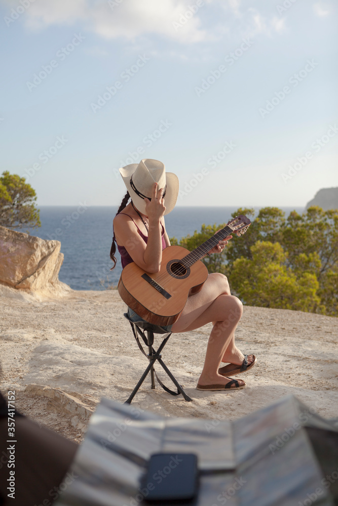 Wanderlust concept of a woman with cowboy look traveling in a van, the girl is wearing a hat and the evening light shines in the sky. Pretty woman play the guitar in seascape of Majorca . Van trip.
