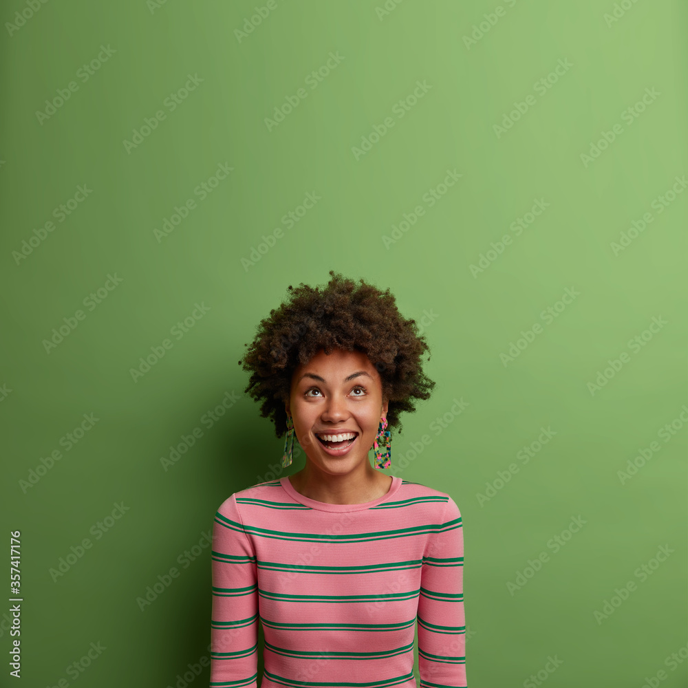 Sociable carefree dark skinned woman looks above with great pleasure and smile, sees amazing offer or promo, new kinds of clothes, has white perfect teeth, isolated on green background. Copy space Sociable carefree dark skinned woman looks above with great pleasure and smile, sees amazing offer or promo, new kinds of clothes, has white perfect teeth, isolated on green background. Copy space