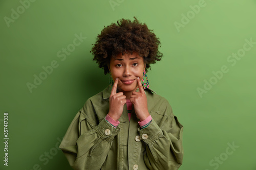 Portrait of dark skinned woman touches fingers near corners of lips, pretends smiling, has unhappy expression, wears green jacket, poses indoor. Unreal smile. People and facial expressions concept